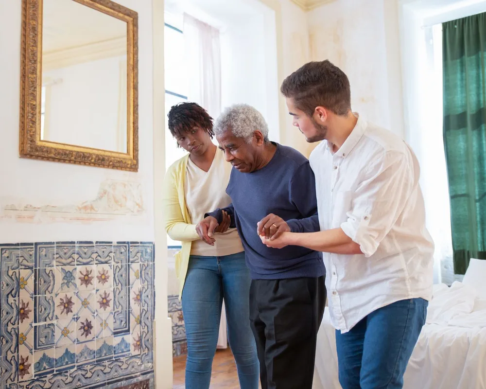 Two clinicians guarding an older man on either side as he walks in his bedroom at home.
